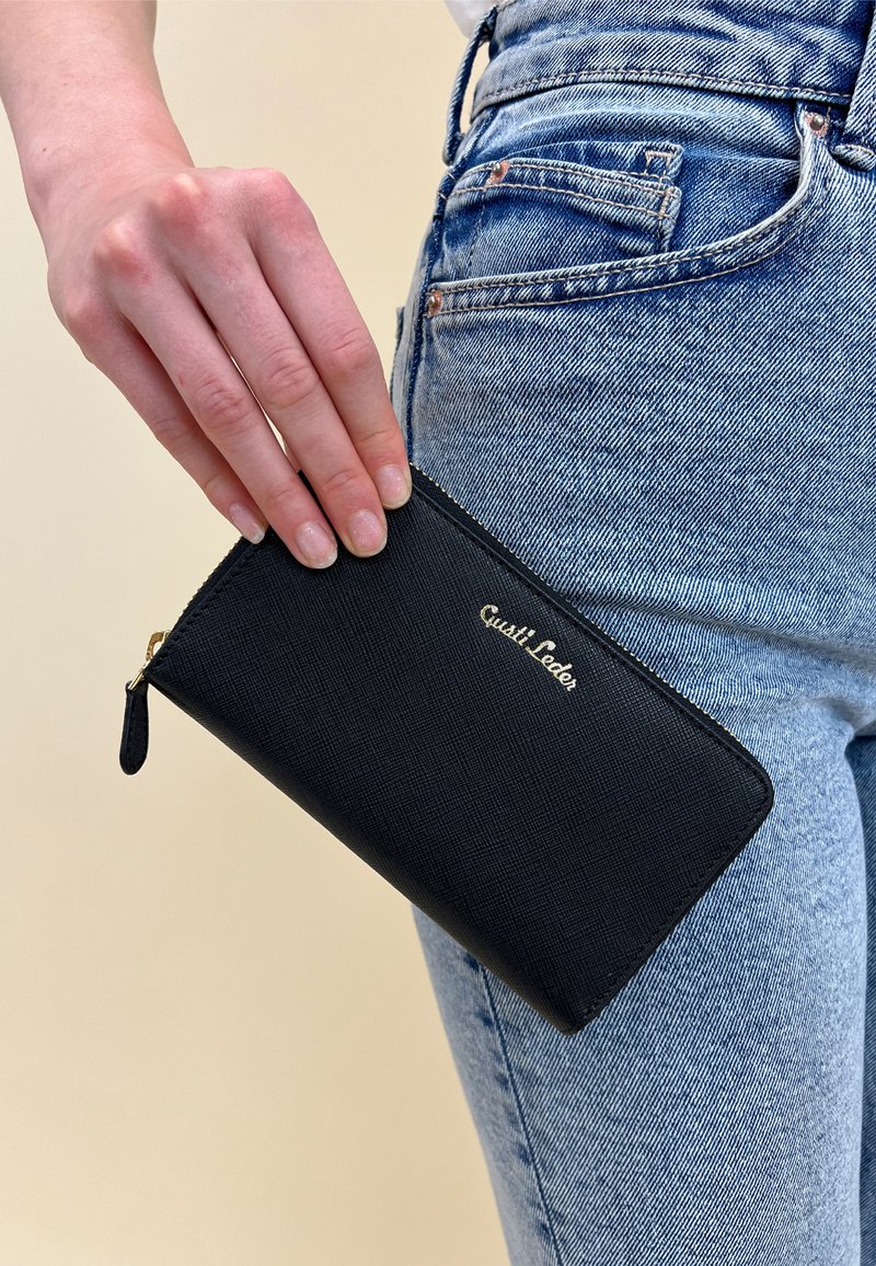 Black zip wallet made of textured material, featuring gold logo, rectangular shape, and a small zip pull tab; held near denim jeans.