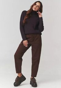 Kobieta z długimi brązowymi włosami, ubrana w czarny sweter z wysokim kołnierzem, ciemne brązowe spodnie i czarne buty na platformie, pozując na jednolitym jasnym tle.