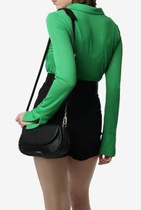 Borsa a tracolla nera con bordatura intrecciata, texture liscia e chiusura in metallo argento. Indossata sopra una blusa verde a maniche lunghe e pantaloni corti neri.