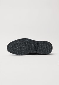 La suola di una scarpa in gomma nera presenta una superficie texturizzata con scanalature e bordi arrotondati. Il design include un'impronta centrale del logo.