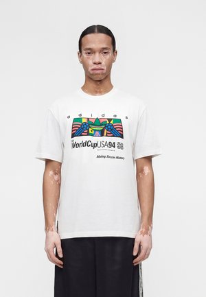 Άτομο με λεύκη που φοράει λευκό μπλουζάκι Adidas World Cup USA 94 και μαύρο παντελόνι, στέκεται μπροστά σε απλό φωτεινό φόντο.