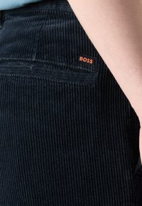 Gros plan de la poche d’un pantalon en velours côtelé noir avec un patch logo BOSS orange et une main posée à côté de la poche.