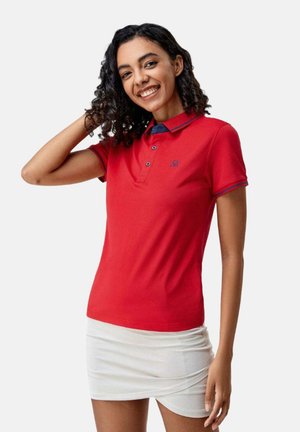 Femme souriante aux cheveux bouclés portant un polo rouge et une jupe blanche, posant avec une main touchant ses cheveux.