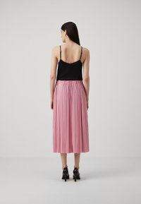 Kvinne som står med ryggen til iført en svart ermeløs topp, rosa plisserte midikjole og svarte høyhælte sko mot en ensfarget bakgrunn.