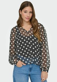 Femme aux longs cheveux bruns portant un blouse noire en voile à pois sur un débardeur noir, accompagnée de jeans bleus, la main dans la poche.