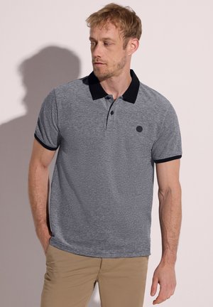 Mężczyzna z jasnymi włosami, ubrany w szarą koszulkę polo z czarnym kołnierzykiem i brzegami rękawów, stoi z jedną ręką w kieszeni beżowych spodni.