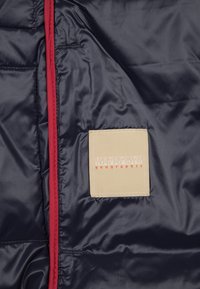 Interior de chaqueta acolchada azul marino con etiqueta cuadrada beige que dice "NAPAPIJRI geographic" y forro de cremallera rojo.