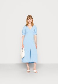 Vestido midi de bolinhas azul claro com mangas bufantes, decote em V, fenda frontal e textura suave; combinado com sandálias de salto brancas e uma bolsa.