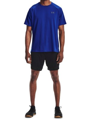 Blaues Sport-T-Shirt mit kurzen Ärmeln, runder Ausschnitt und Logo. Kombiniert mit schwarzen Shorts und schwarzen Sportschuhen.
