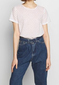 Biała koszulka z krótkim rękawem z małym kwiatowym wzorem w kolorze czerwonym i zielonym, zestawiona z wysokimi niebieskimi dżinsami z okrągłą klamrą pasa.