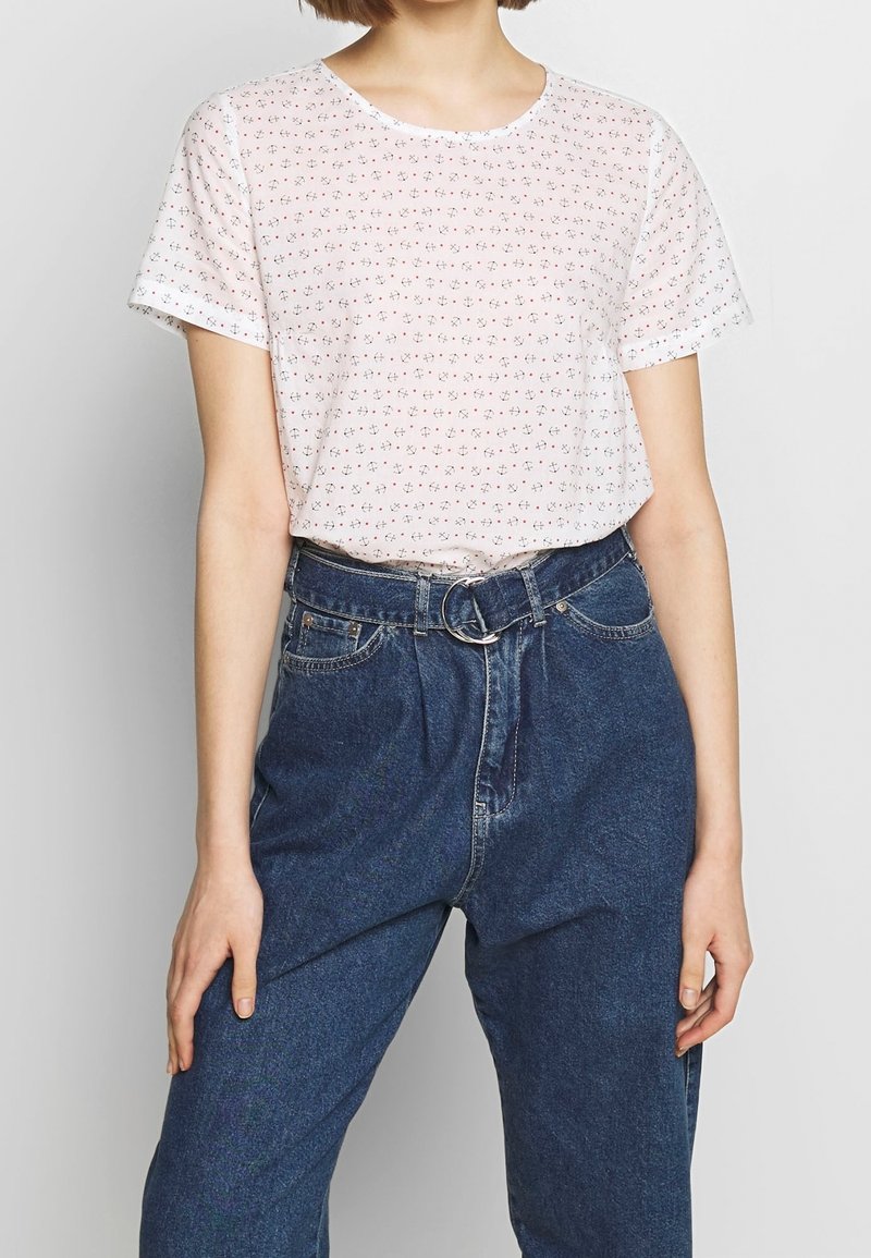 Biała koszulka z krótkim rękawem z małym kwiatowym wzorem w kolorze czerwonym i zielonym, zestawiona z wysokimi niebieskimi dżinsami z okrągłą klamrą pasa.