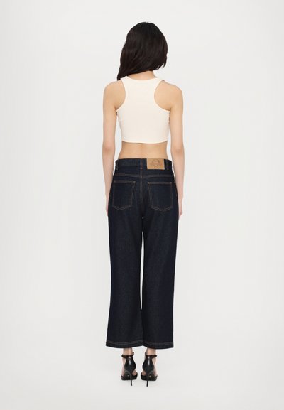 Jeans in denim scuro a vita alta con gambe ampie, dotati di una toppa in pelle beige sul retro. Abbinati a un top bianco aderente senza maniche.