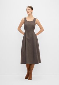 Robe sans manches marron avec un corps ajusté et une jupe évasée, faite de tissu texturé ; associée à des bottes marron montantes.