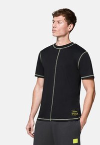 Czarna koszulka z krótkim rękawem z kontrastującym zielonym szwem, okrągłym dekoltem i akcentem logo blisko dolnej krawędzi. Gładka tekstura materiału.