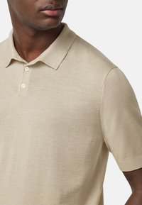Beige polo tričko vyrobené z lehké pleteniny, s límečkem a dvěma knoflíky, přiléhavým střihem a krátkými rukávy.