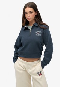 Sudadera recortada azul marino con cuello con cremallera, logo de "Superdry Athletics", puños acanalados, combinada con pantalones de chándal color crema con detalles del logo.