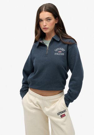 Granatowy, krótki polar z zamkiem przy kołnierzu, z logo "Superdry Athletics", z prążkowanymi mankietami, noszony z kremowymi spodniami dresowymi z detalem logo.