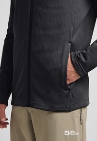 Schwarze Zip-Jacke mit strukturiertem Muster, ausgestattet mit Seitentaschen und elastischen Bündchen. Kombiniert mit hellbeigen Hosen mit Branding.