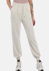 Pantaloni sportivi di colore beige chiaro con vestibilità rilassata, cintura elastica e due tasche anteriori. Realizzati in un tessuto morbido con polsini elastici alle caviglie.