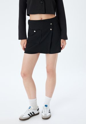 Falda envolvente negra con un bolsillo frontal, detalles de botones plateados y costuras en contraste. Combinada con zapatillas blancas y calcetines.