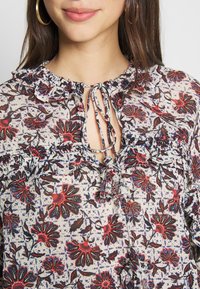 Blouse florale en tissu léger et transparent couleur crème, ornée de motifs de fleurs rouges et bleues, avec un col froncé et une fermeture à nouer.