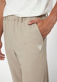 Šviesiai rudos spalvos sportinės kelnės su tekstūruotu audiniu, elastingu juosmeniu ir šonine kišene, kurioje yra baltas trikampio formos logotipas.
