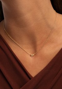 Goldene Halskette mit einer zarten Kette, die zwei kugelförmige Akzente und einen kleinen Diamanten aufweist, getragen auf einem nackten Hals vor einem braunen Stoff.