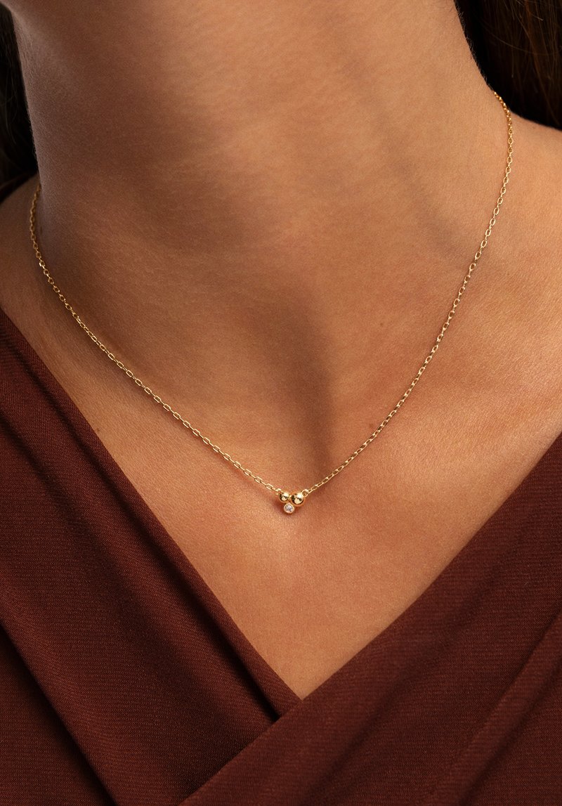 Goldene Halskette mit einer zarten Kette, die zwei kugelförmige Akzente und einen kleinen Diamanten aufweist, getragen auf einem nackten Hals vor einem braunen Stoff.