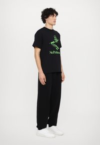 Sort bomulds T-shirt med grøn grafisk tryk og teksten "No Problem" på fronten, parret med sorte bukser og hvide sneakers.
