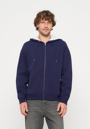 Jeune homme aux cheveux bouclés portant un sweat à capuche bleu marine zippé et un jean gris, debout devant un fond uni clair.