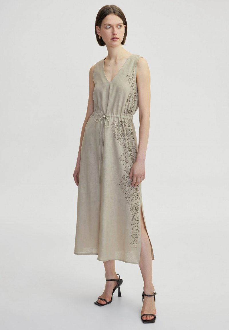 Abito senza maniche beige con scollo a V, vita elastica e fessure laterali; presenta un design testurizzato su un lato. Indossato con sandali neri con tacco alto.