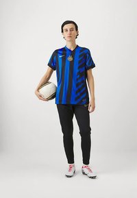 Henkilö mustassa ja sinivalkoraidallisessa jalkapallopaidassa, mustissa housuissa, valkoisissa kengissä pitelee valkoista ja kultasta jalkapalloa tasaisella taustalla.