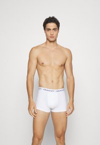 Boxeri din bumbac alb cu un tiv de culoare bleumarin, având o potrivire strânsă și o textură netedă. Modelul este fără tricou și stă în picioare.