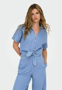 Heled sinine teksashirt lühikeste varrukatega, nööbiga esiosa ja seotava vöökohtaga. Sile tekstuur, millel on mugav ja lõdvestunud istuvus.