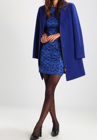 Sininen villakangastakki, jossa on läppä, istuvan sinisen pitsimekon päällä. Läpinäkyvät mustat pilkkuverkkosukat ja mustat kiiltonahkaiset korkokengät viimeistelevät asun.