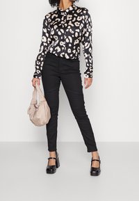 Žena v černých kalhotách s prošitím na kolenou, černobílé leopardí vzorované blůze, černých botách Mary Jane a držící béžovou kabelku.