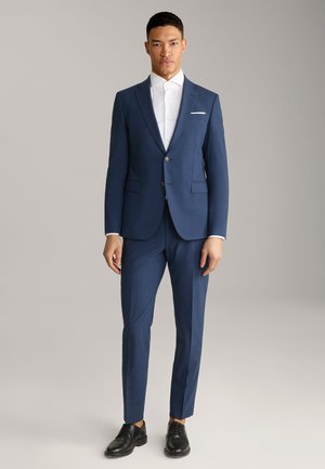 Costume bleu marine en tissu texturé avec une coupe ajustée, comprenant une chemise blanche et des chaussures en cuir noir. Fermeture à deux boutons.