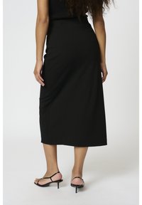 Jupe noire mi-longue avec une texture lisse, taille ajustée et drapés subtils. Associée à des sandales à talons nus noires.