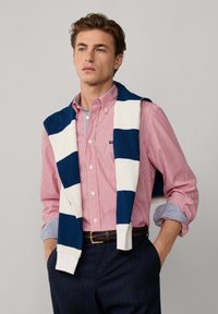 Rot und weiß gestreiftes Hemd mit Knöpfen, kombiniert mit einem blau und weiß gestreiften Pullover, der über die Schultern gelegt ist. Dunkel gestreifte Hose.