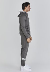 Šedý športový overal s kapucňou, ktorý má na nohaviciach biely logovaný pás. Biele tenisky dopĺňajú outfit. Hladká textúra, priliehavý strih.