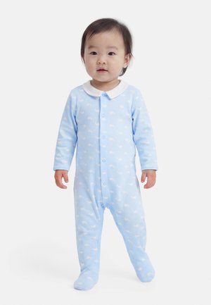 Småbarn står iført en lyseblå heldragt med fødder og et hvidt hvalemønster samt hvid krave på ensartet hvid baggrund.