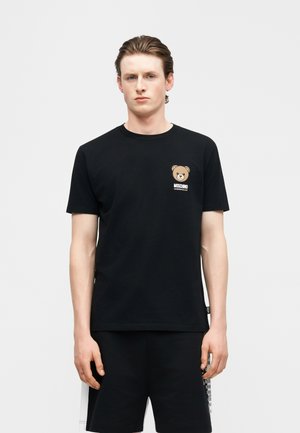 Młody mężczyzna w czarnej koszulce Moschino z małym logo misia na piersi oraz dopasowanych czarnych szortach.