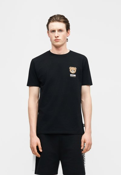 Νεαρός άνδρας που φοράει μαύρο T-shirt Moschino με μικρό λογότυπο αρκουδάκι στο στήθος και αντίστοιχα μαύρα σορτς.