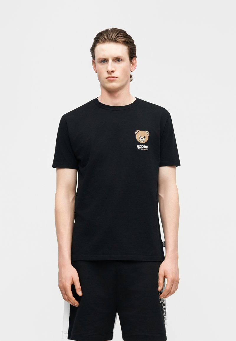 Νεαρός άνδρας που φοράει μαύρο T-shirt Moschino με μικρό λογότυπο αρκουδάκι στο στήθος και αντίστοιχα μαύρα σορτς.
