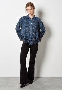 Camisa de denim azul con un patrón texturizado, con un dobladillo de encaje ondulado, cierre de botones y mangas largas, combinada con pantalones acampanados negros.
