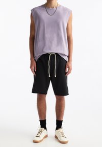 T-shirt violette sans manches à coupe décontractée, associé à un short noir avec cordon de serrage. Chaussures blanches avec des accents beiges et chaussettes noires.