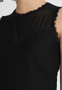 Haut sans manches noir avec un panneau transparent sur le haut de la poitrine et une finition en dentelle le long de l'encolure et des emmanchures, porté par une personne.