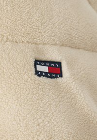 Kremowy polarowy materiał z prostokątną etykietą w czarno-białe wzory z czerwonymi akcentami, na której widnieje napis "TOMMY JEANS." Widoczna jest miękka tekstura.