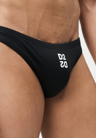 Κοντινή εικόνα της κάτω περιοχής του κορμού ενός ατόμου που φοράει μαύρα μαγιό τύπου σλιπ με λευκό λογότυπο "D2" στη δεξιά πλευρά.