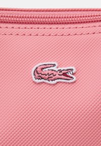 Un tissu texturé rose présente un logo de crocodile brodé en rose clair et noir avec des accents rouges sur la bouche.